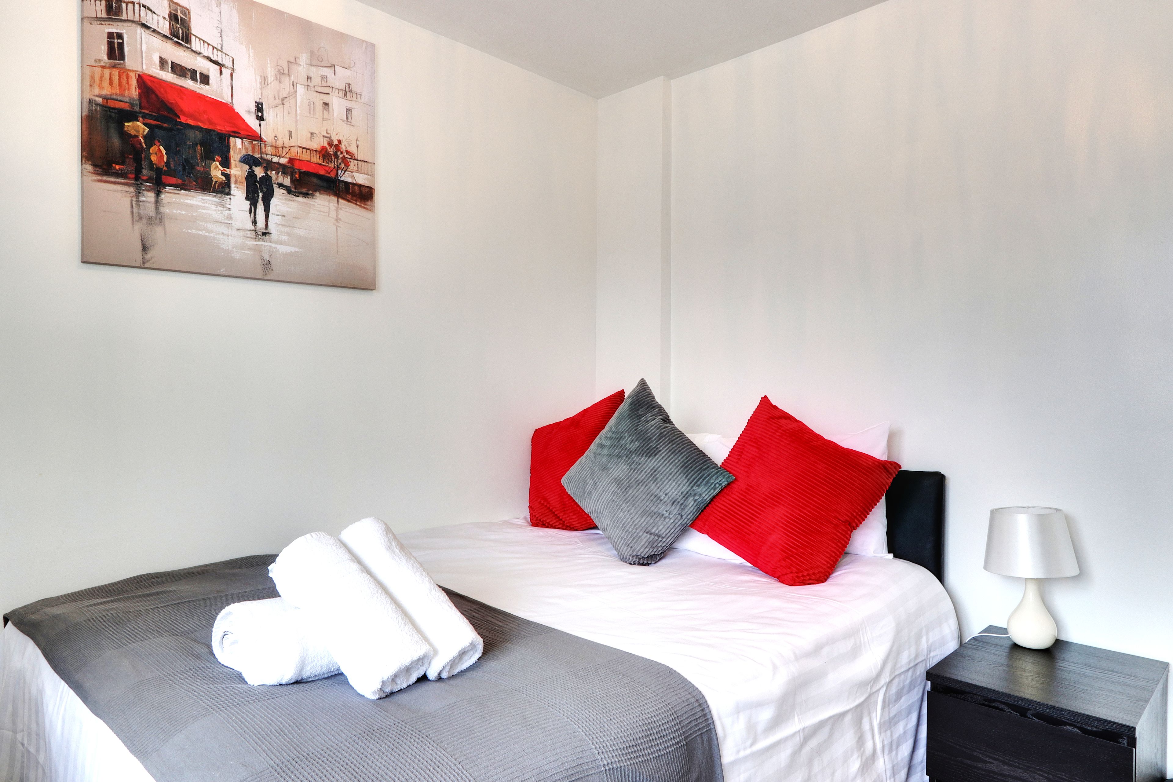 A hotel room with a bed, pillows, a nightstand, a lamp, and a painting on the wall.