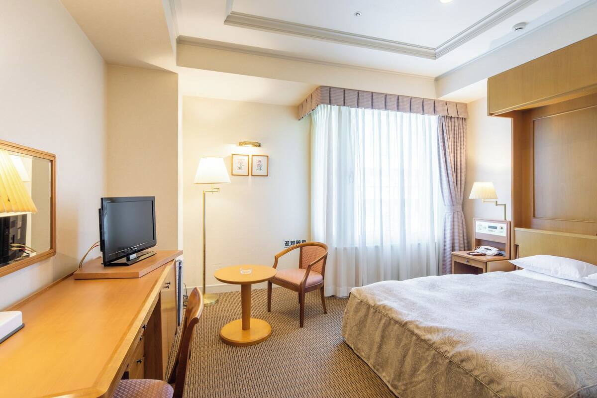 Une chambre d’hôtel comprenant un lit, un bureau avec une télévision, une chaise, une petite table, une lampe et une table de chevet avec un téléphone.