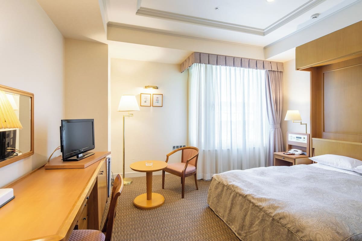 A hotel room with a bed, a desk with a TV, a chair, a small table, a lamp, and a nightstand with a phone.