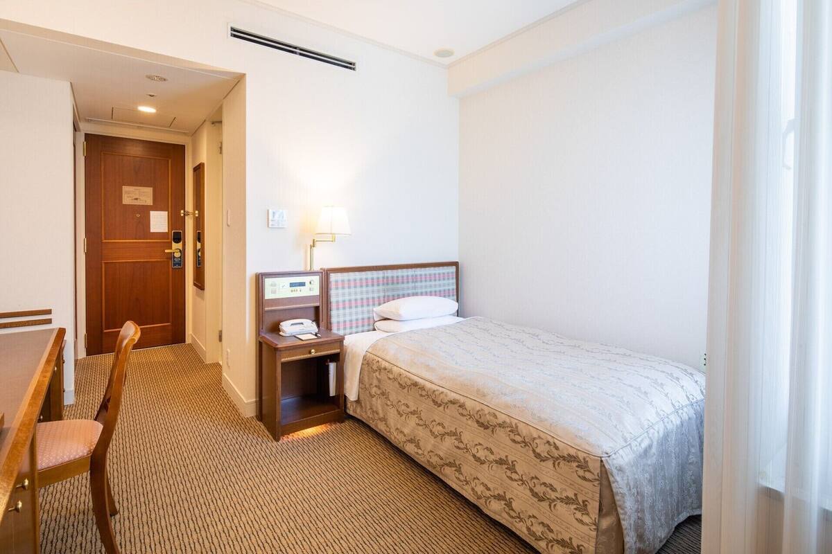 A hotel room with a bed, a desk, a chair, a nightstand, and a lamp.