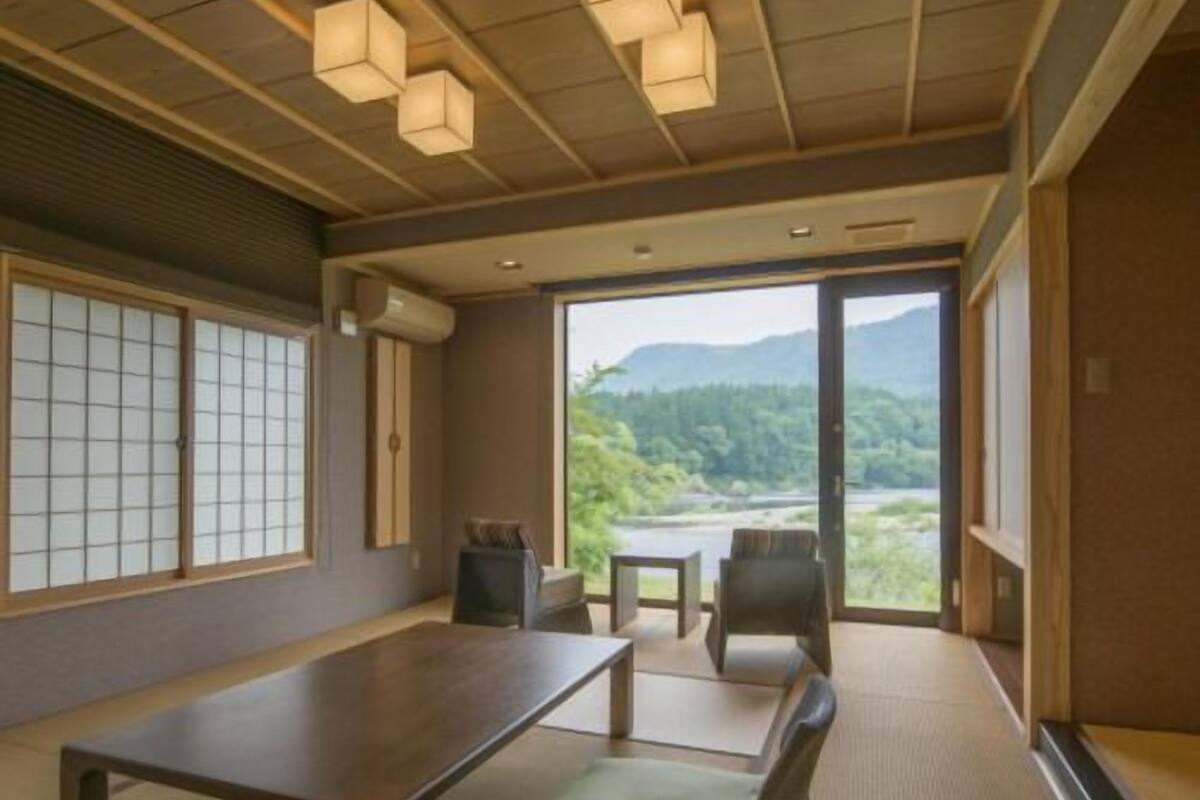 A room with tatami flooring, a low table, chairs, and a view of a mountainous landscape through large windows.