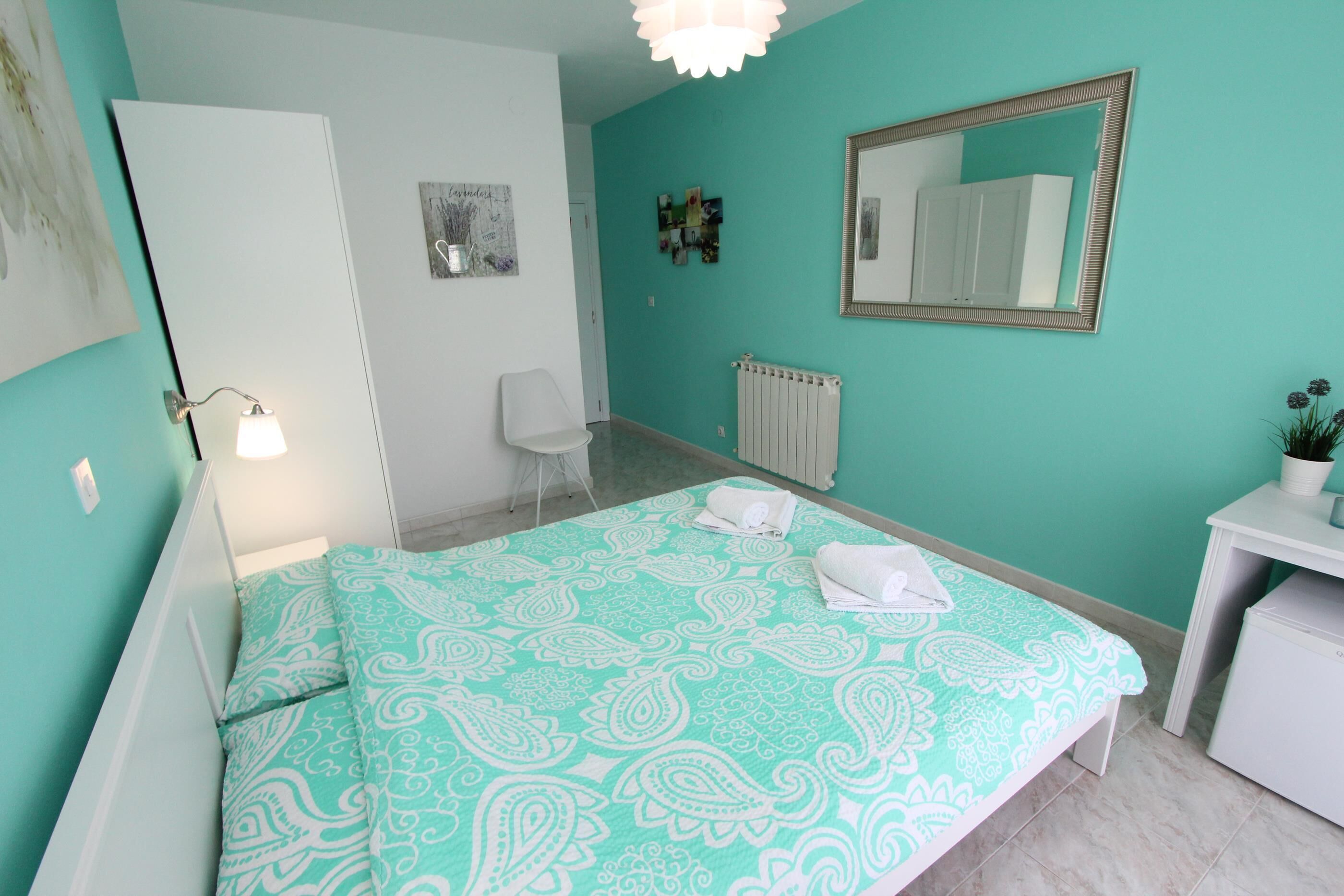 A bedroom with a bed, a desk, a chair, a radiator, and a mirror on the wall.