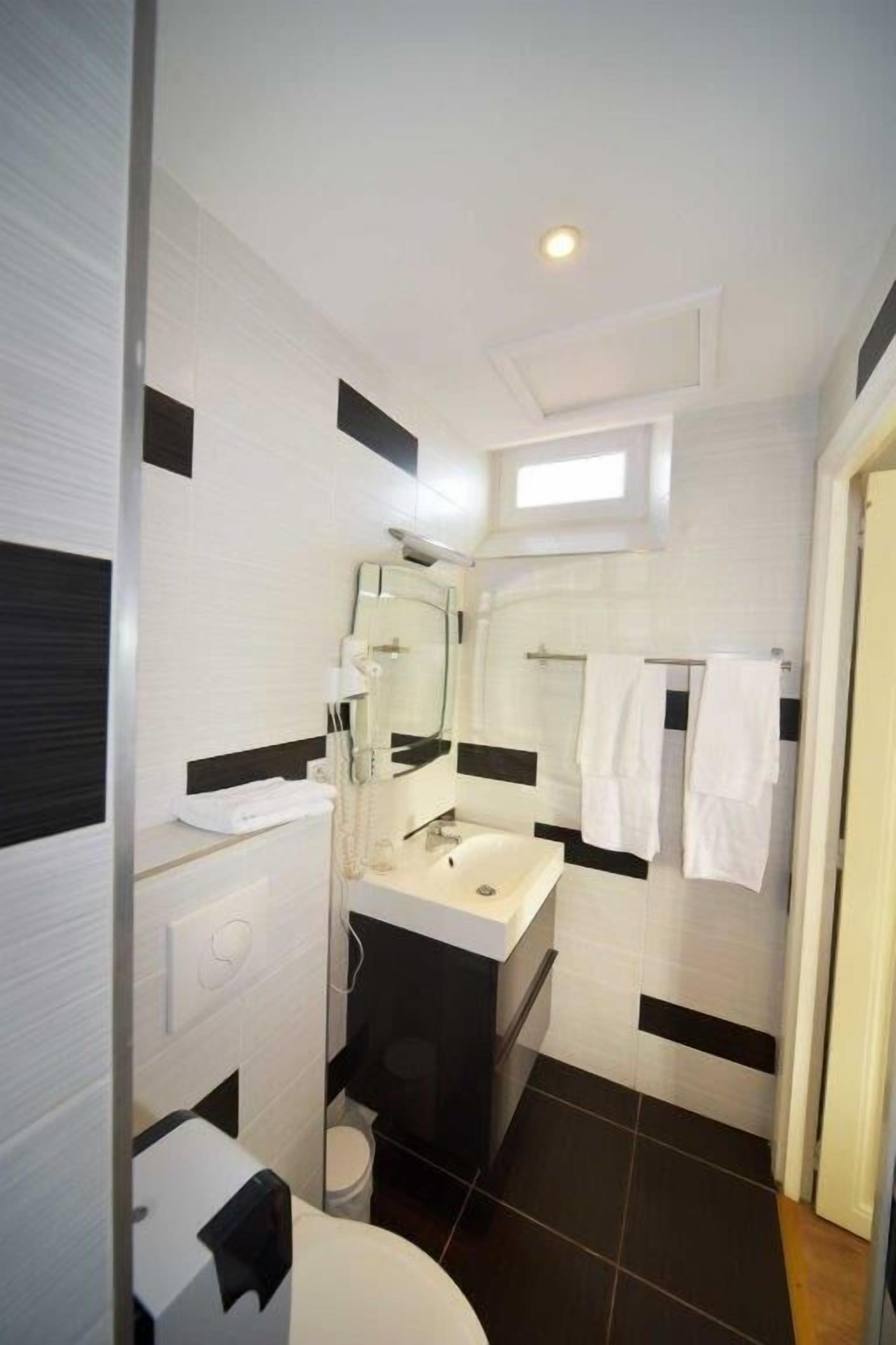 Une petite salle de bain avec un lavabo blanc, un miroir et des toilettes. On y trouve un porte-serviettes sur lequel sont suspendues des serviettes, ainsi qu’une petite fenêtre au-dessus du miroir.