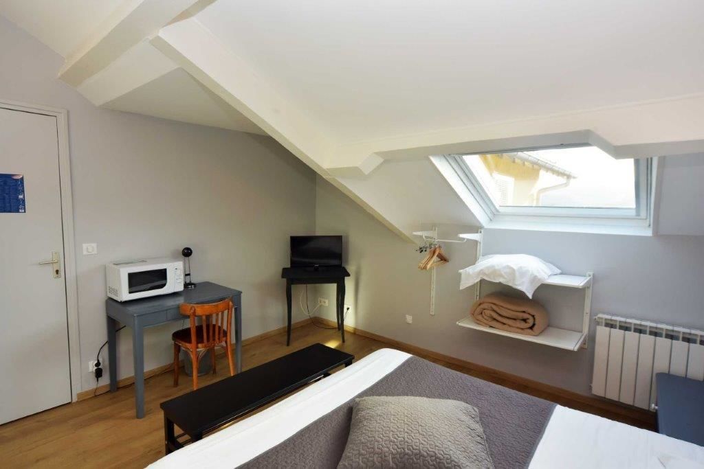 Een kleine, goed verlichte kamer met een schuine kap, een bed, een bureau met een computer, een stoel, een magnetron en een klein raam.