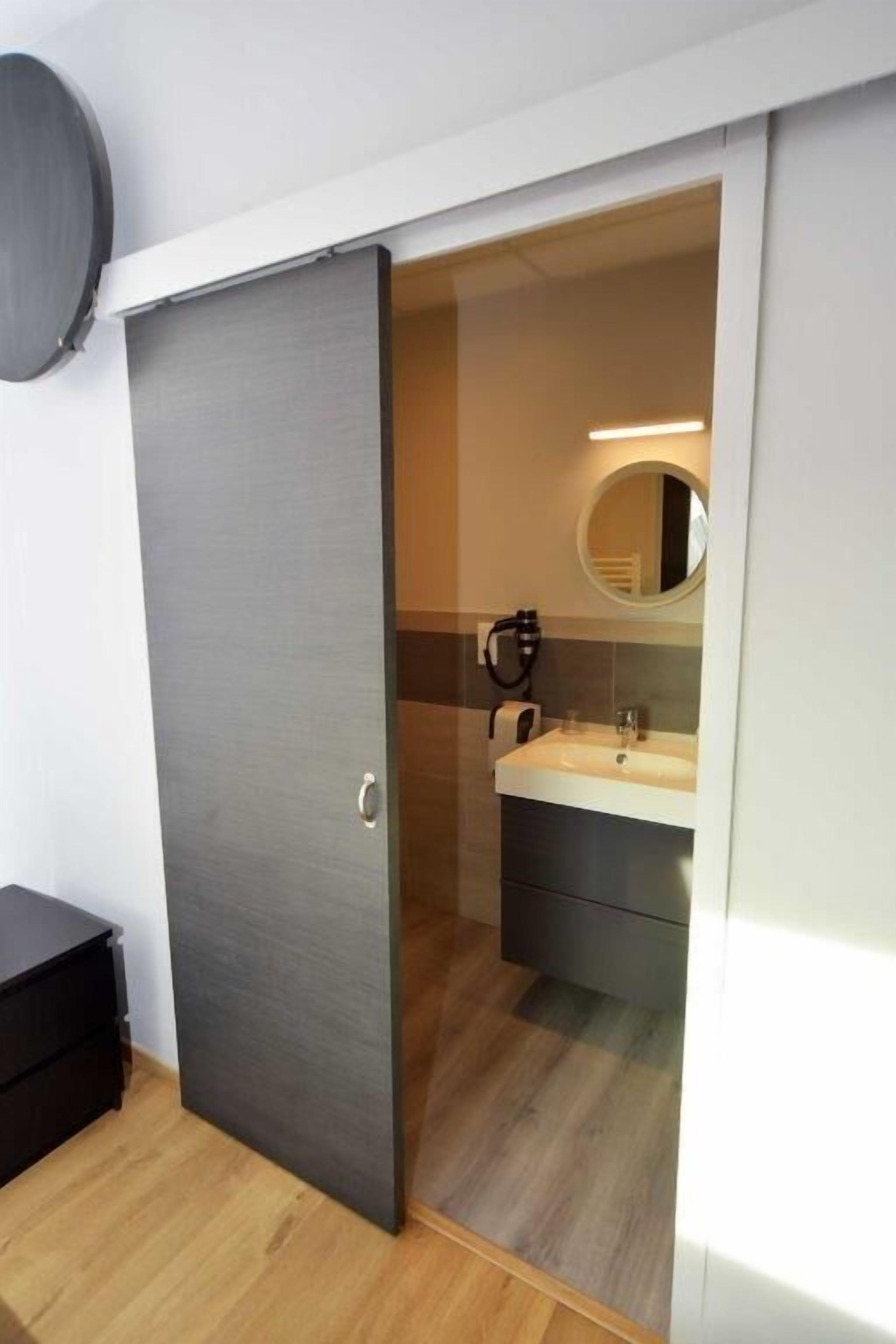 Un baño moderno con una puerta gris, un espejo redondo y un lavabo blanco.