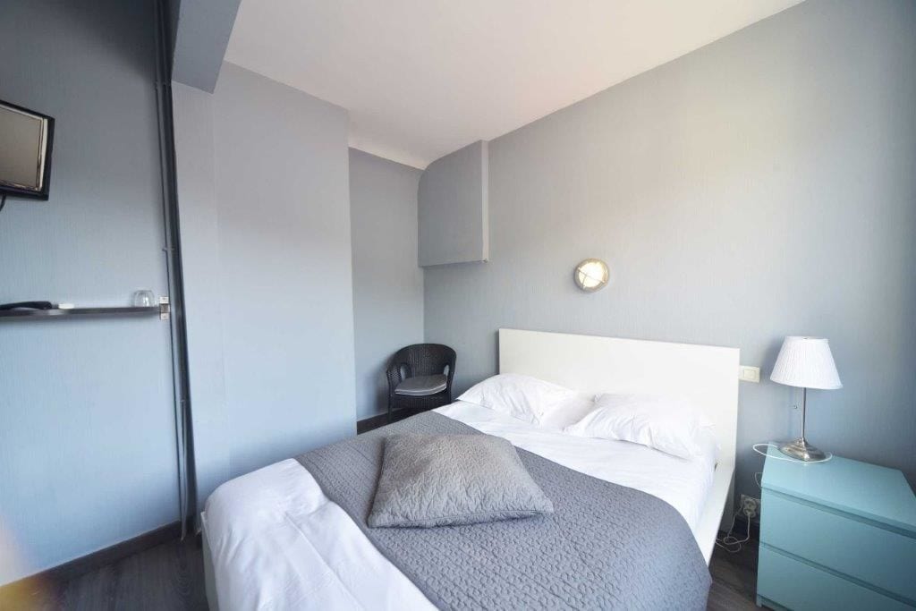 A hotel room with a bed, a chair, a bedside table with a lamp, and a wall clock.