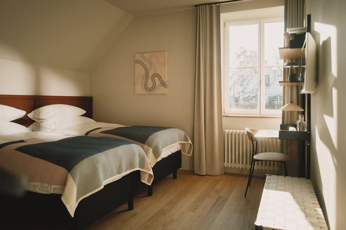 A hotel room with two beds, a window with curtains, a radiator, a table with a chair, and a wall-mounted shelf with items.