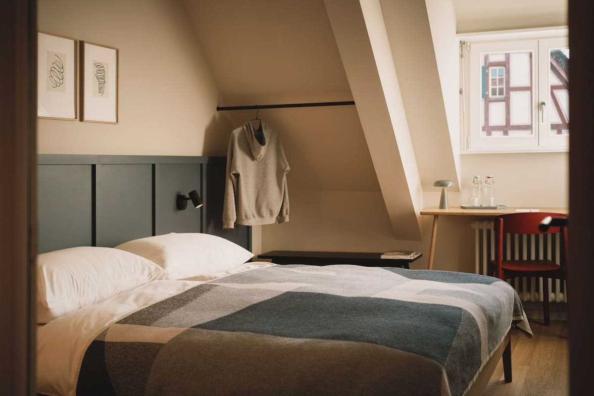A bedroom with a bed, a desk, and a chair. There is a window with a view of buildings outside.
