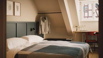 Un dormitorio con cama, escritorio y silla. Se ve una ventana con vistas a edificios.