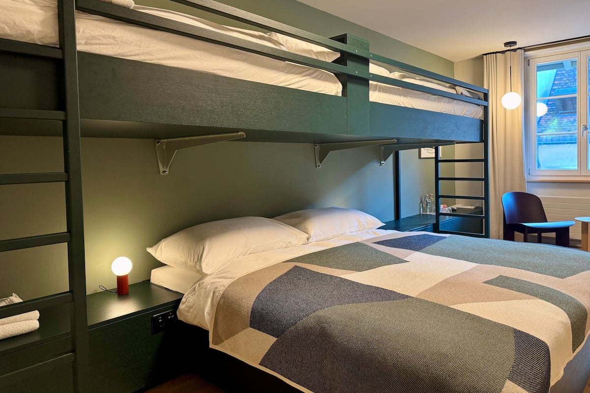 A bunk bed with a quilted bedspread, a bedside table with a lamp, and a chair near a window.
