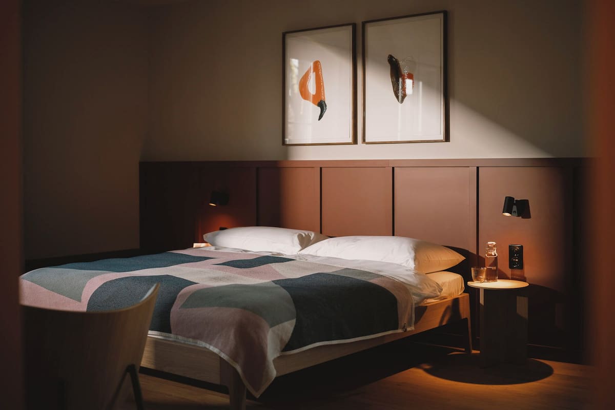 A hotel room with a bed, a chair, a bedside table with a lamp, and two framed pictures on the wall.