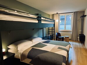 A bunk bed with a geometric-patterned bedspread, a small table with a chair, and a window with curtains. - Landgasthof (Riehen)