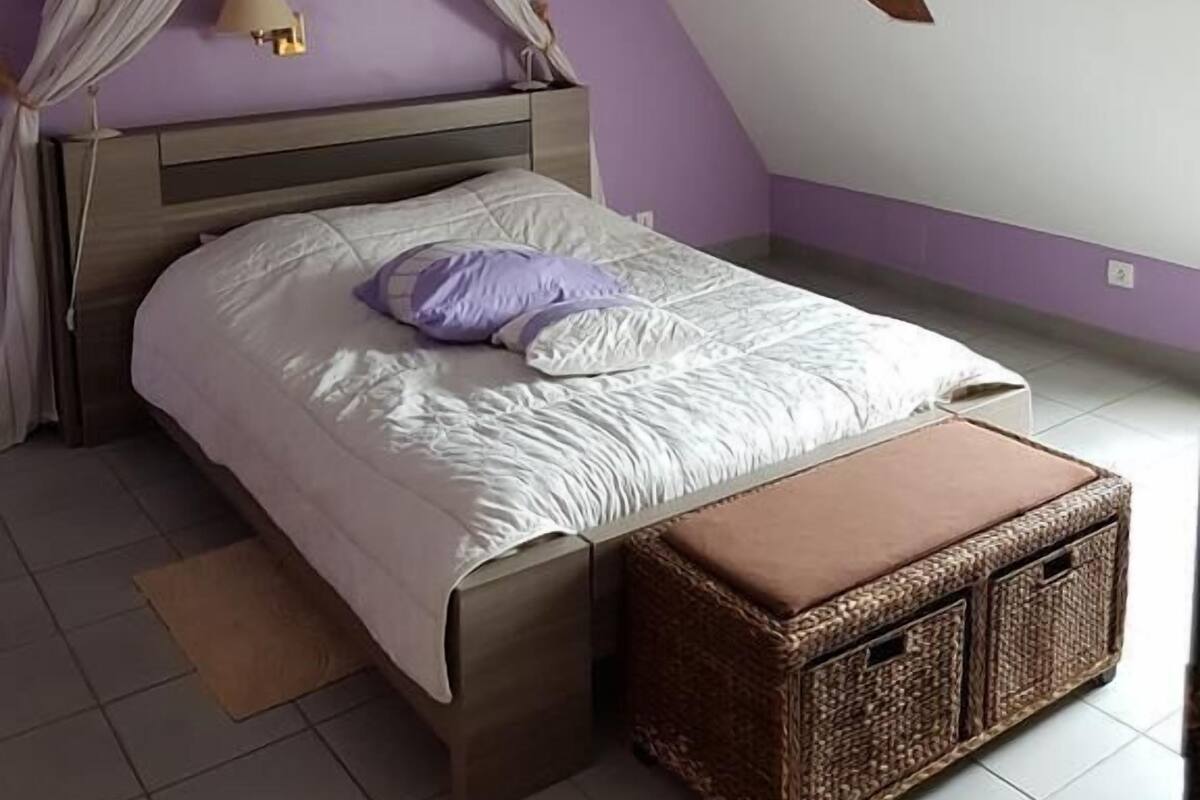 Een slaapkamer met een bed, nachtkastje, rieten kist en een plafond met zichtbare balken.