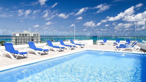 A rooftop pool with blue and white lounge chairs, a clear sky, and a view of the ocean.