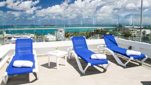 A rooftop lounge with three blue sunbeds and a small table with a drink, overlooking a beach and ocean.