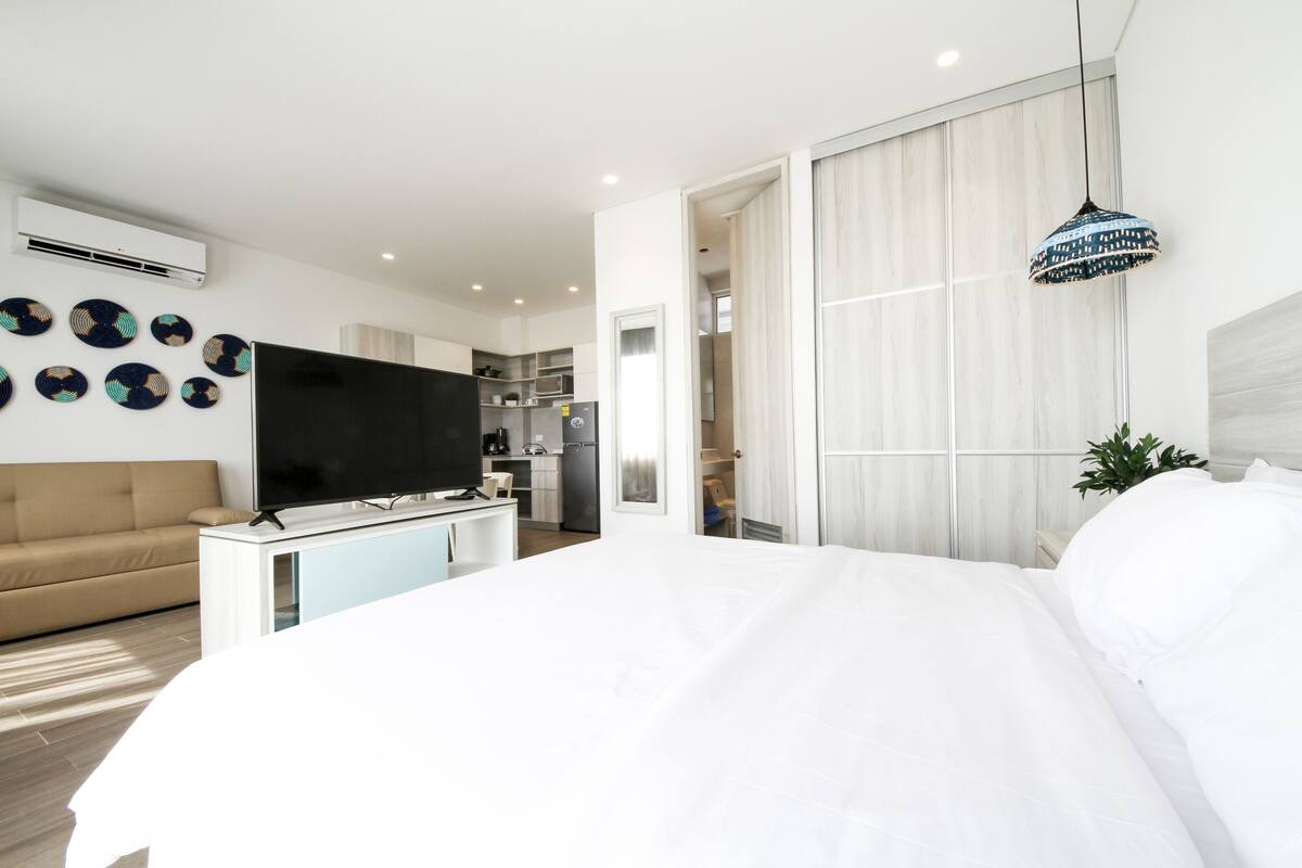 A modern hotel room with a large bed, a flat-screen TV, a sofa, and a kitchenette in the background.