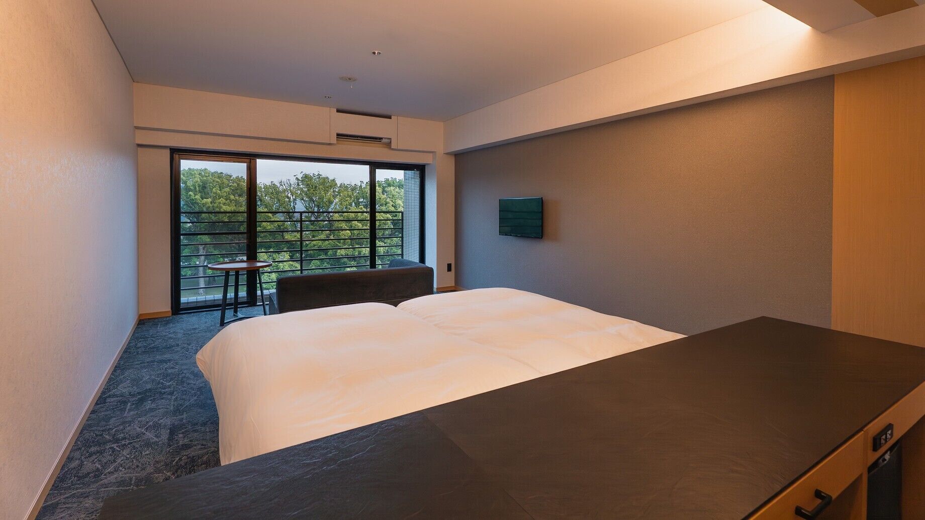 Hotelkamer met een bed, een klein tafeltje, een stoel, een televisie en een balkon met uitzicht op bomen.