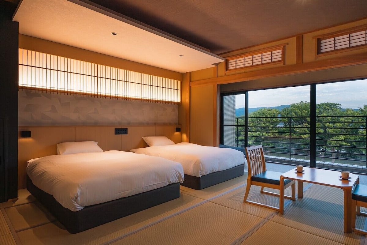 A hotel room with two beds, a small table, chairs, and a large window offering a view of trees and mountains.
