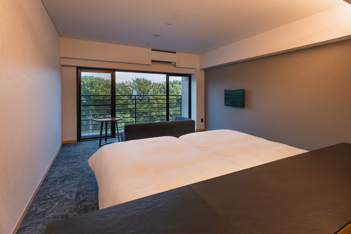 A hotel room with a bed, a small table, a chair, a TV, and a balcony with a view of trees.