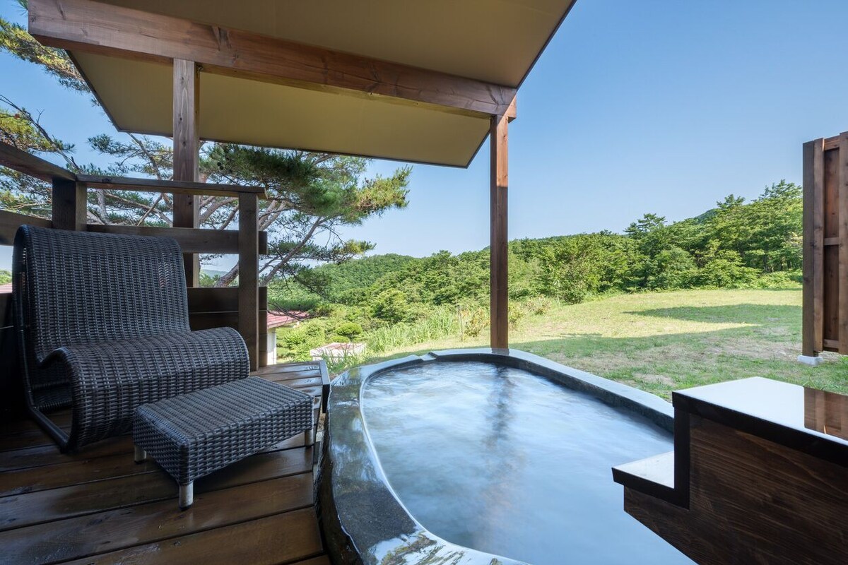 A hot tub with a wooden deck and seating area, surrounded by greenery and a clear sky.