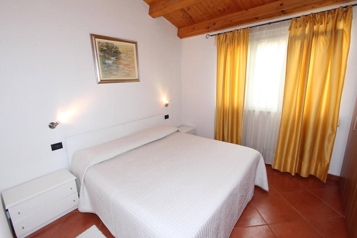 Quarto com cama, mesa de cabeceira, luminária na parede, janela com cortinas e um quadro na parede.