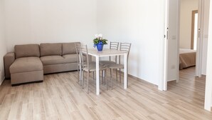 A living room with a beige sofa, a dining table with four chairs, and a blue vase with flowers. - Aria di Vacanza - S'Abba Lughente Trilo (Olbia)