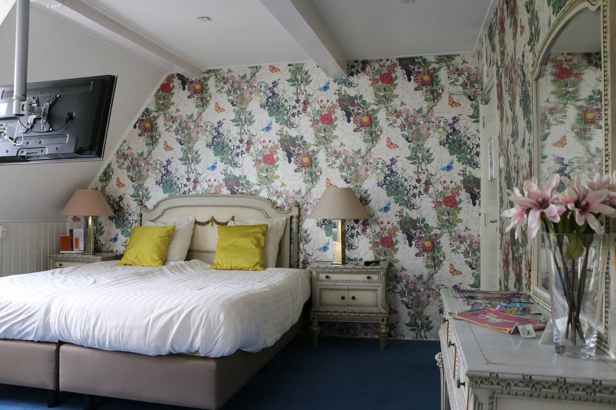 Un dormitorio con cama, mesitas de noche, cómoda, espejo y papel tapiz con motivos florales.