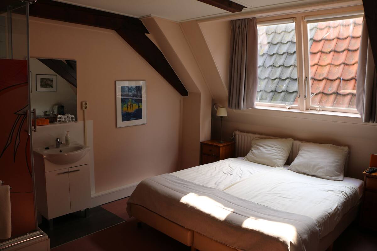 Un dormitorio con cama, una mesita de noche, una ventana con cortinas, un radiador, un cuadro en la pared y un baño con lavabo y espejo.