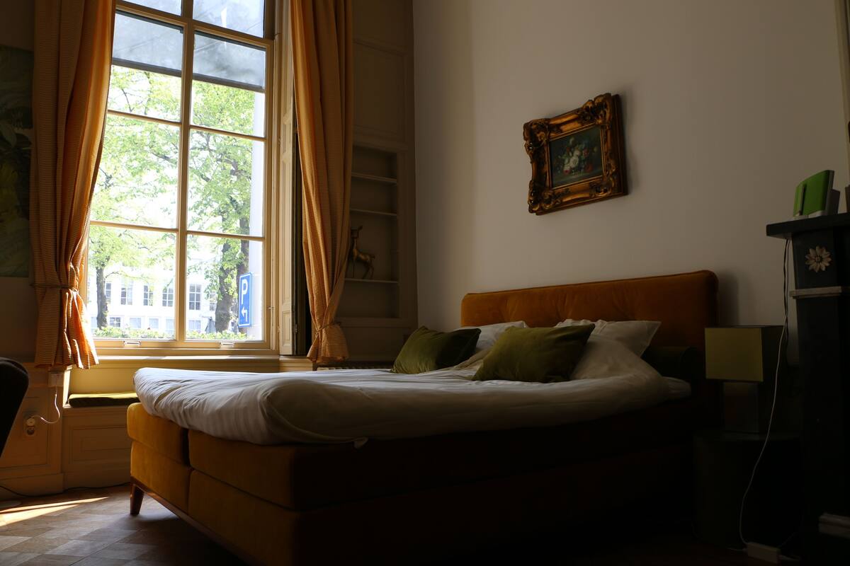Un dormitorio con una cama, una ventana con cortinas, un cuadro enmarcado en la pared y una mesita de noche con una lámpara.