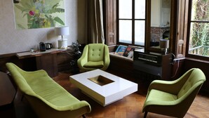 Een kamer met een bureau, twee groene fauteuils, een witte salontafel en een groot raam met gordijnen.