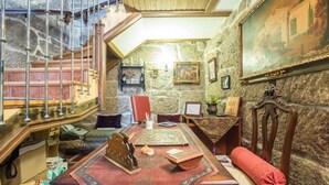 Une pièce avec un mur en pierre, un escalier en bois, une table recouverte d’une nappe rouge et une chaise avec un coussin rouge.