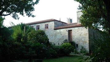 Un edificio de piedra con techo de tejas rojas, rodeado de abundante vegetación y un pequeño jardín.