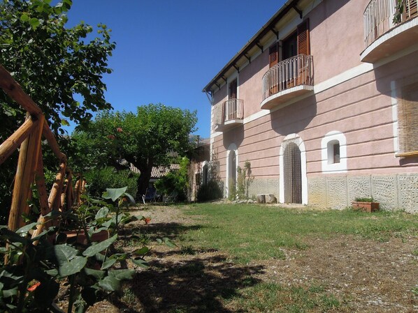 A building with a pink facade, a balcony with a metal railing, and a small garden area with grass and plants.