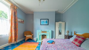 A bedroom with a patterned bedspread, a fireplace, a wicker chair, and a dresser. - Stag and Hen Friendly House (Bath)