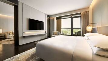 A modern hotel room with a large bed, a flat-screen TV mounted on the wall, a seating area with chairs, and a large window offering a view of the outdoors.