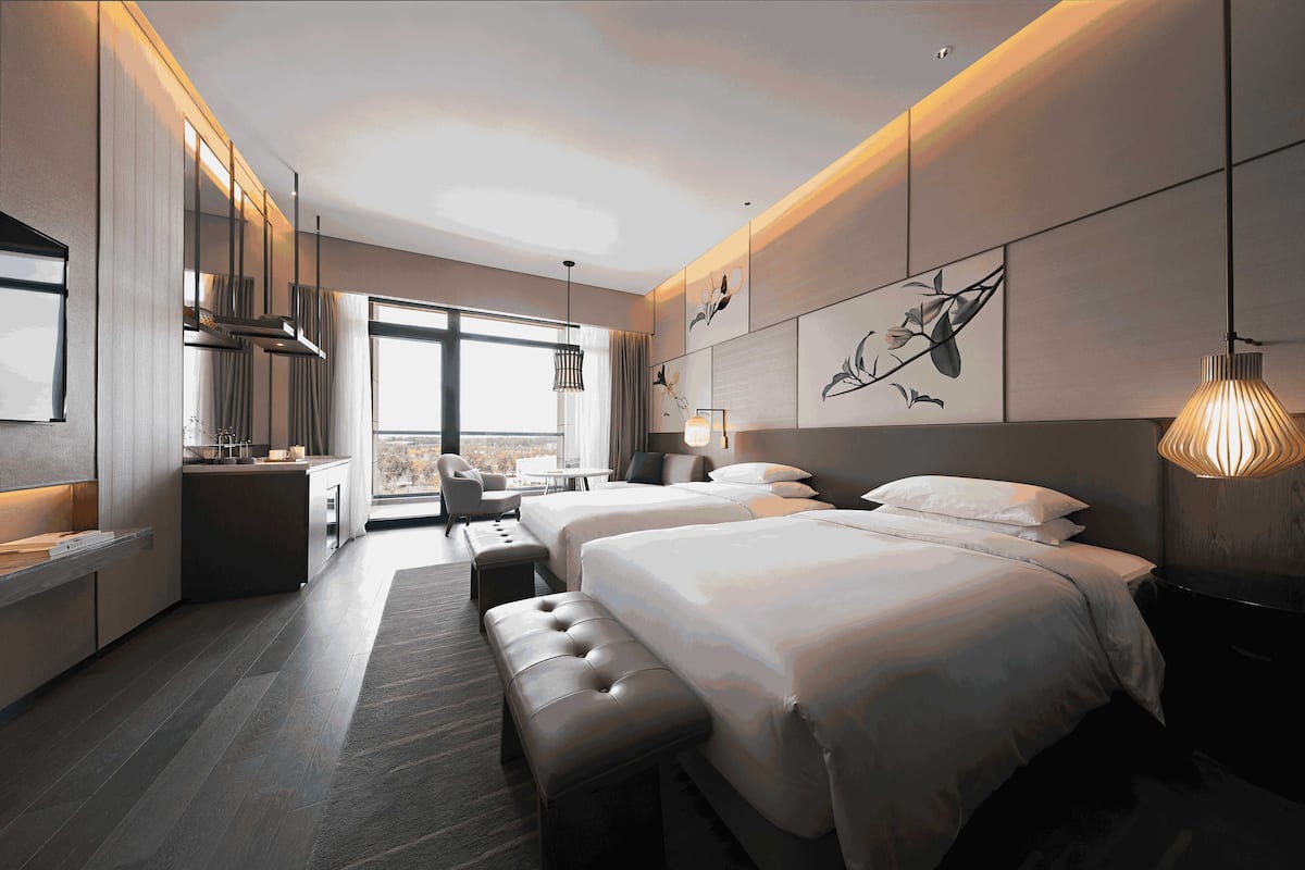 A modern hotel room with two beds, a sitting area, and a view of the city.