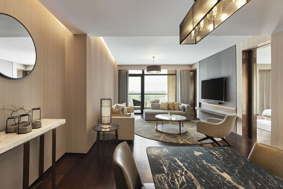 A modern hotel room with a large window, a flat-screen TV, a sofa, a coffee table, and a dining table.