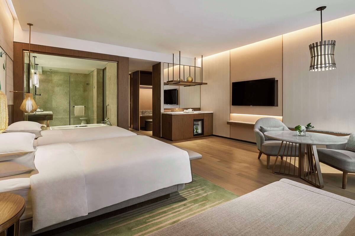 A modern hotel room with a large bed, a sitting area with a table and chairs, a TV mounted on the wall, and a bathroom with a glass shower enclosure.