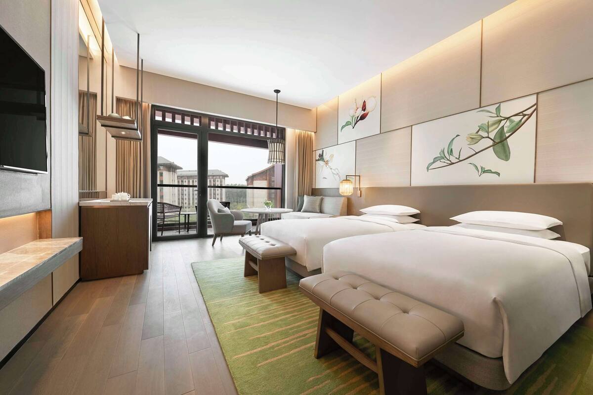 A modern hotel room with two beds, a balcony, a TV, and artwork on the walls.
