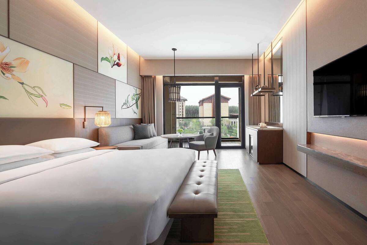 A modern hotel room with a large bed, a sofa, a dining area, and a balcony with a view.