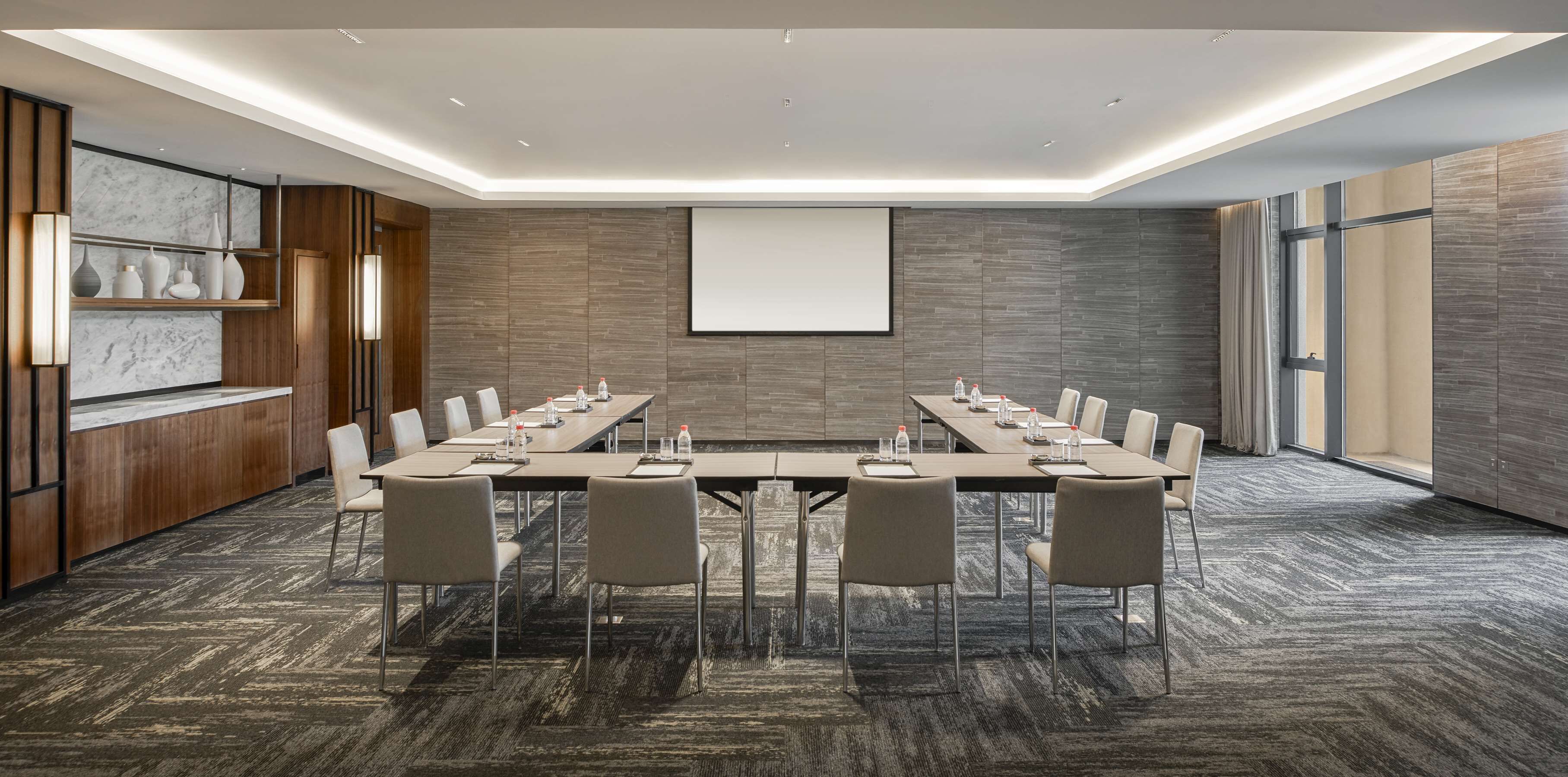 Une salle de conférence avec une longue table, des chaises, un écran de projection et un mur en marbre doté d’étagères.