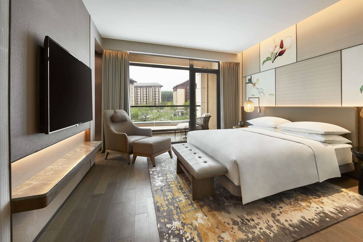 A modern hotel room with a large bed, a flat-screen TV, a comfortable armchair, and a view of the cityscape through large windows.