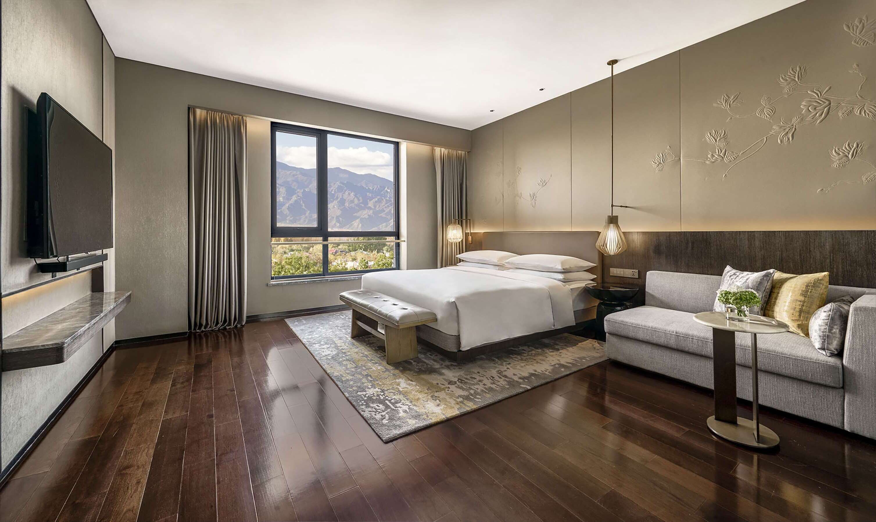 A modern hotel room with a large bed, a sofa, a TV, and a view of mountains.