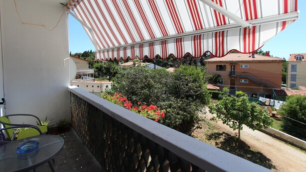 Un balcón con toldo a rayas, una mesa con superficie de vidrio y vista a una zona residencial con edificios y árboles.