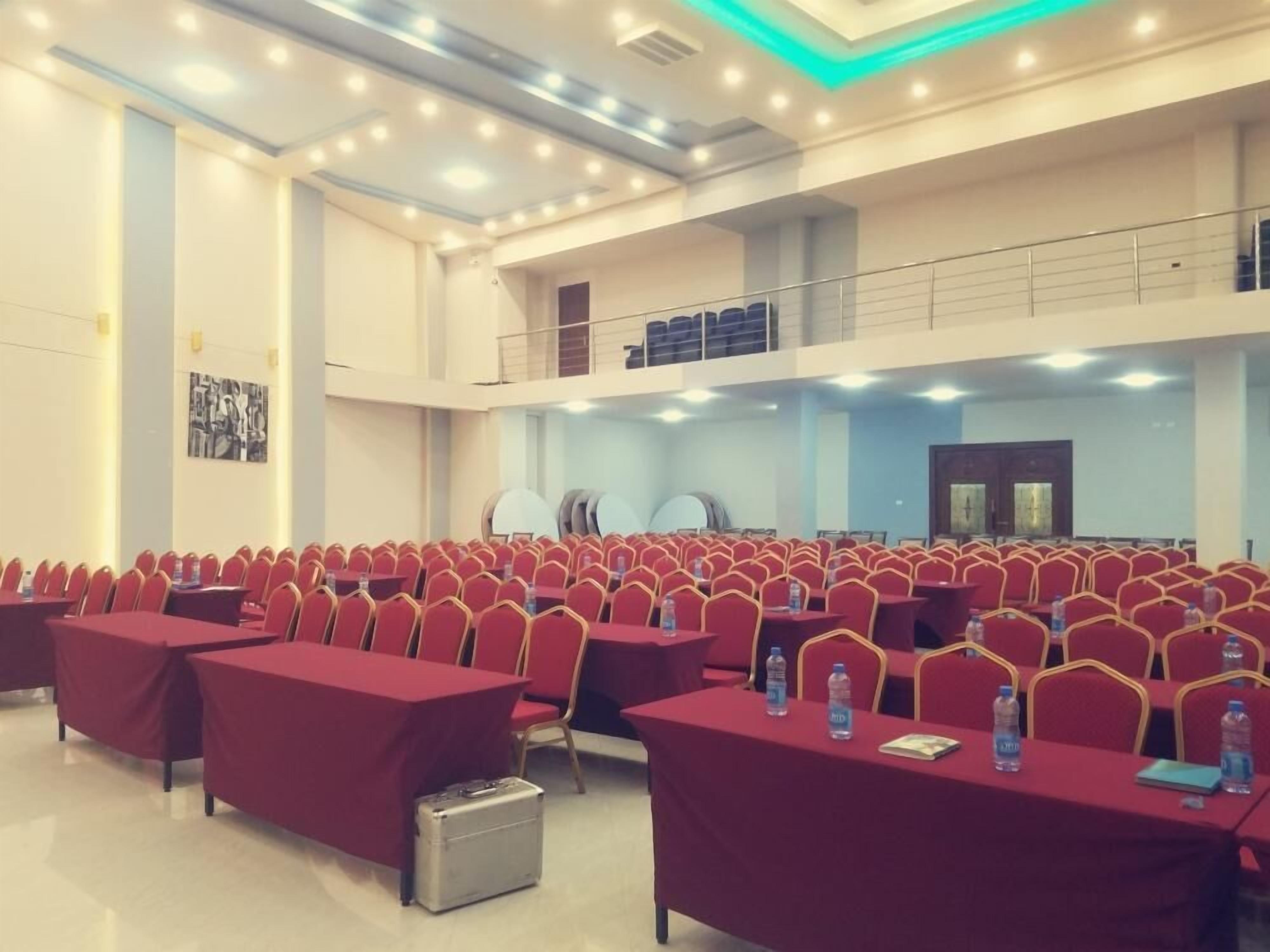 Un amplio salón de conferencias con filas de sillas rojas y mesas, equipado con botellas de agua y una maleta.