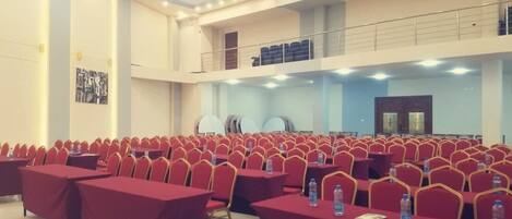 Un amplio salón de conferencias con filas de sillas rojas y mesas, equipado con botellas de agua y una maleta.
