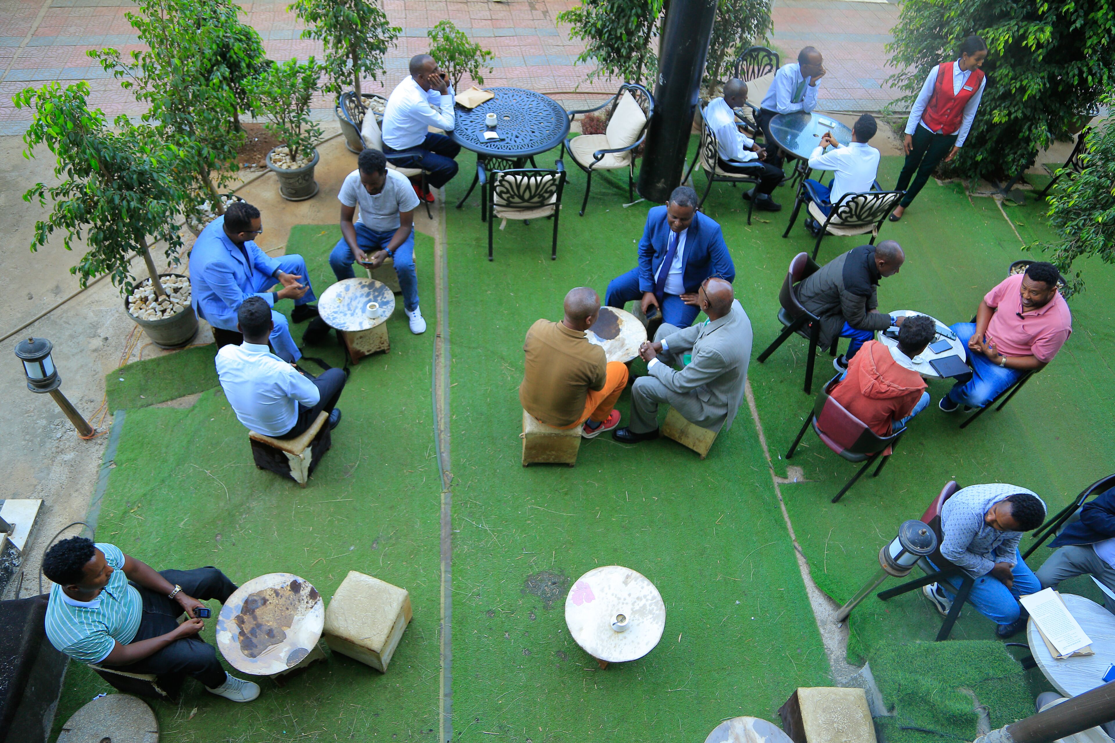 A group of people seated around a round table in an outdoor setting with green artificial turf, surrounded by potted plants and outdoor furniture.