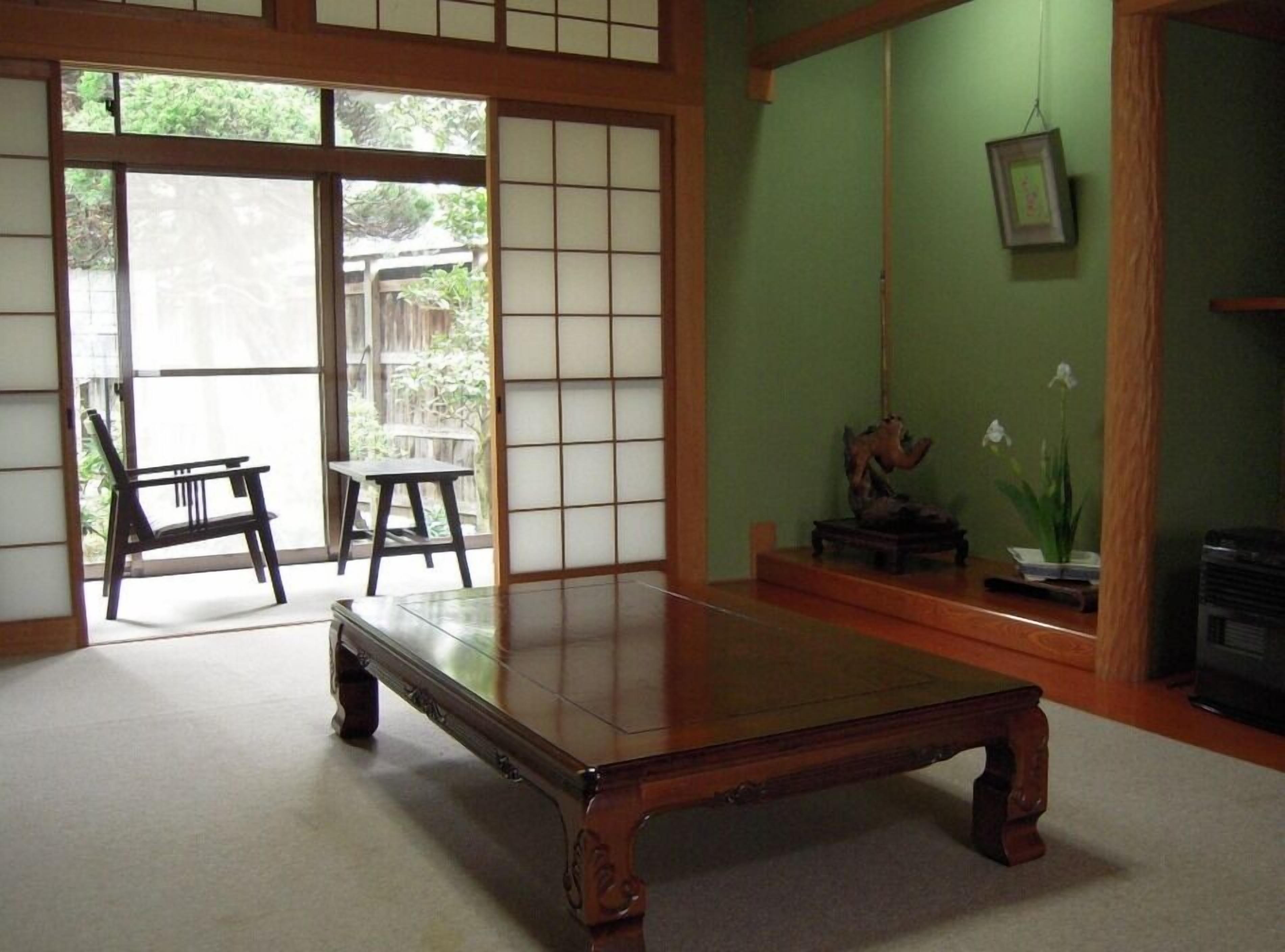 A traditional Japanese room with tatami flooring, a low wooden table, a sliding door leading to a garden, and a green wall with a framed picture.