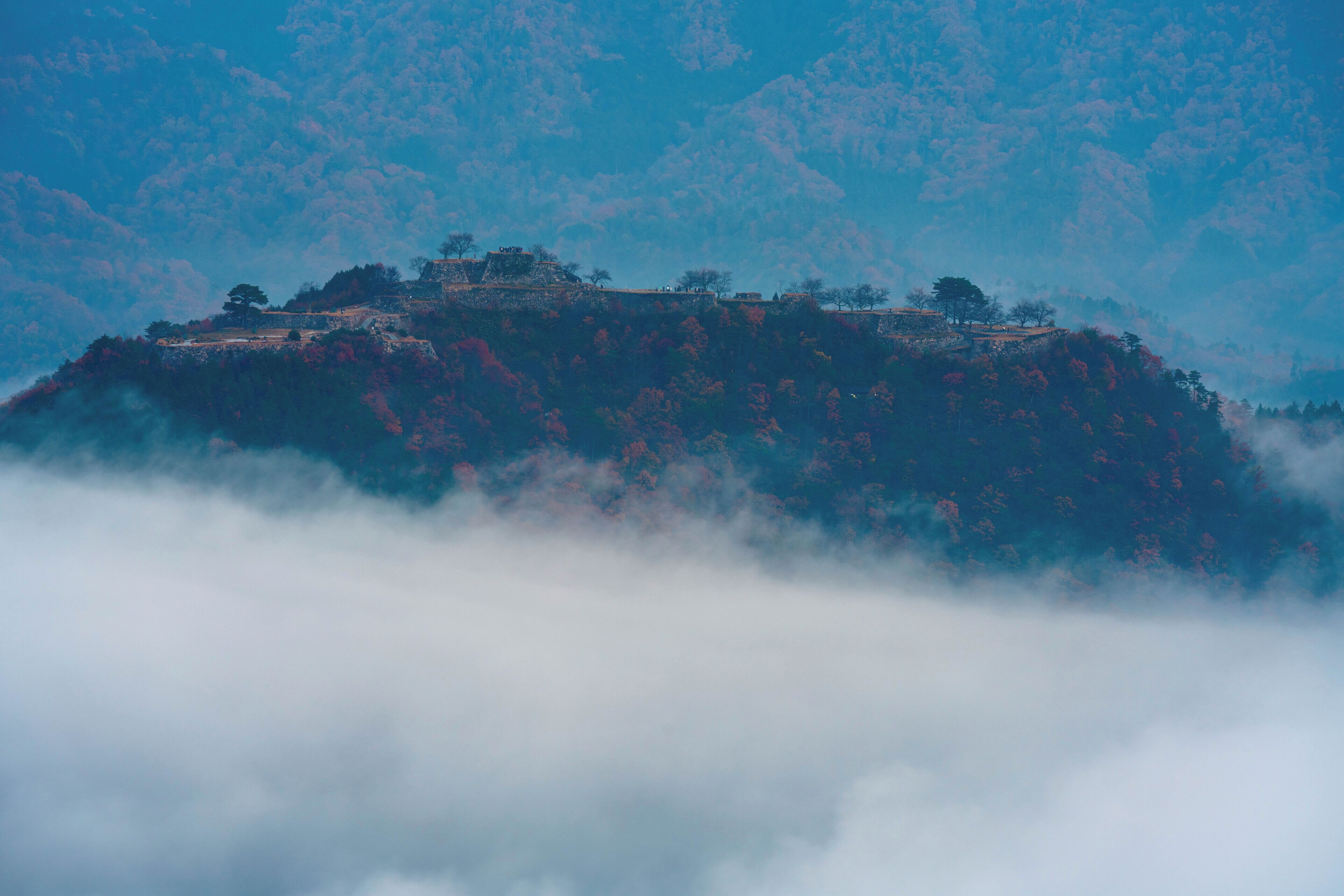 A mountainous landscape with a building on top, surrounded by trees and clouds.