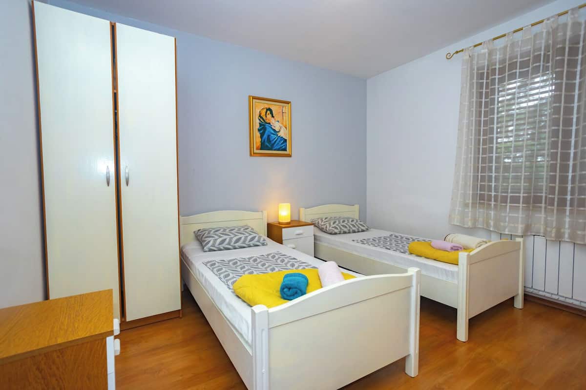 A bedroom with two single beds, a wardrobe, a nightstand with a lamp, and a window with a sheer curtain.